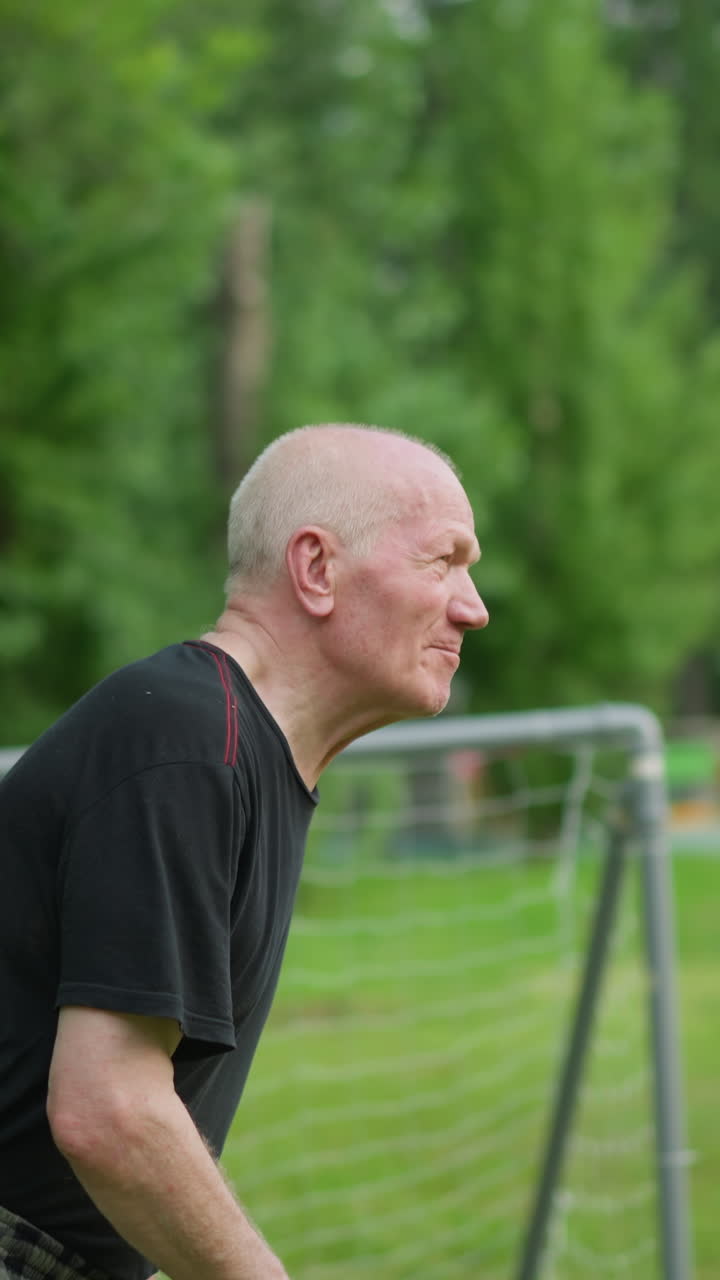 An elderly man skillfully heads a soccer ball away, focusing intently with a blurred view of a goalpost, trees, and a building in the background
