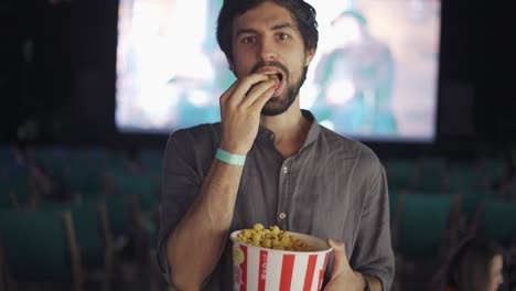 A-bearded-guy-eats-popcorn-at-the-summer-cinema,-smiling-to-the-camera