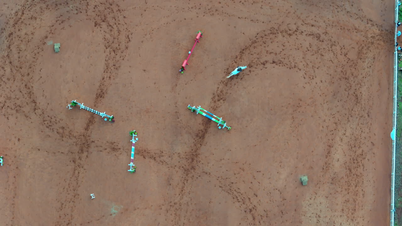 Overhead drone view of a reddish equestrian training arena showing horse track marks and jumping fences arranged in rows, likely for show jumping practice, real time, drone tracking shot