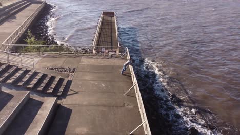 Aerial-orbit-shot-of-young-man-standing-on-railing-in-front-of-river-shore-during-golden-sunset-in-Buenos-Aires