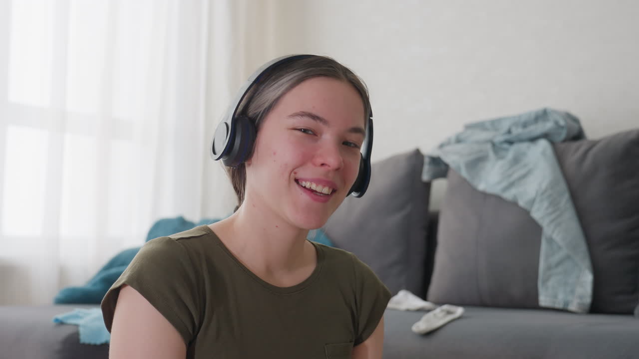 Smiling young woman wearing headphones seated indoors enjoying sound of music, natural light streaming through curtains illuminating relaxed atmosphere, soft expression