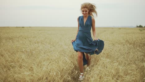 Hübsche-Junge-Frau-In-Einem-Langen-Blauen-Kleid,-Die-Durch-Ein-Goldenes-Weizenfeld-Rennt-Und-In-Die-Kamera-Blickt.-Freiheitskonzept.-Zeitlupe