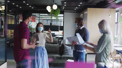 Diverse-business-people-wearing-masks-standing-using-laptop-and-goign-through-paperwork-in-office