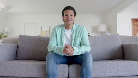 portrait of happy biracial man sitting on sofa smiling in living room, copy space, slow motion