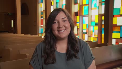 a pretty young ethnic female smiles brightly at the camera inside a church sanctuary while sitting in a pew