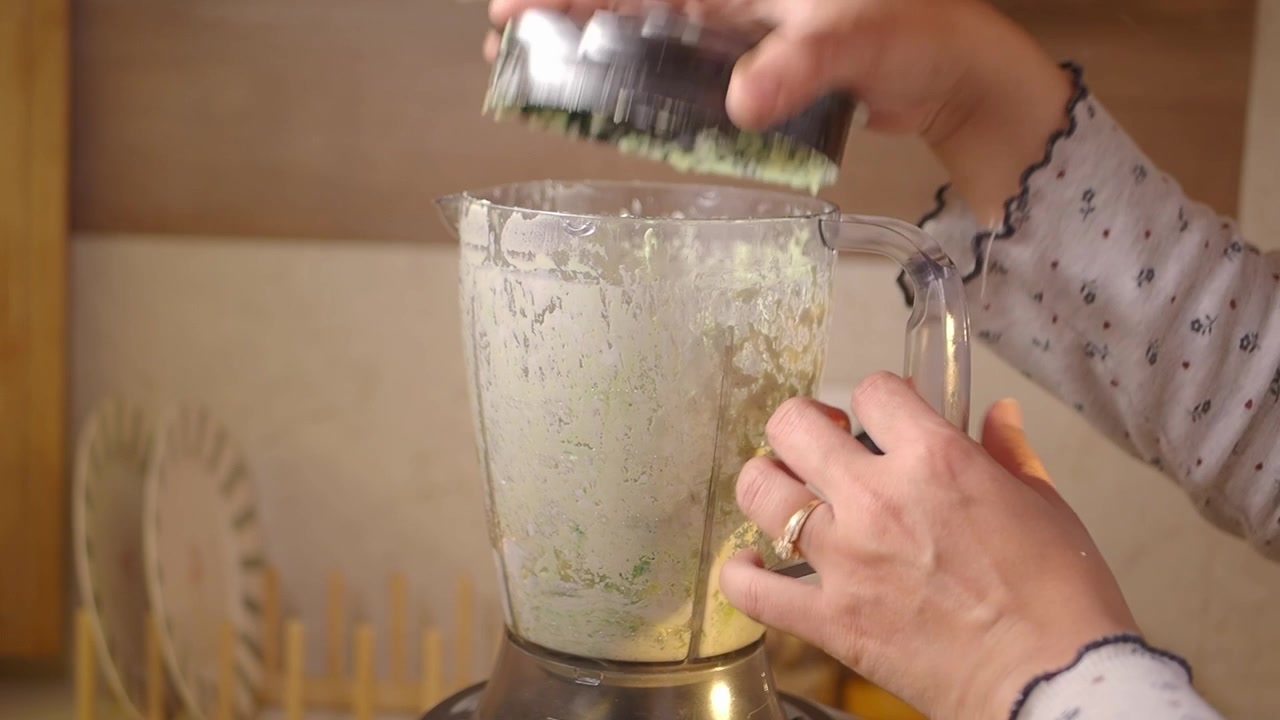 Donna che apre il frullatore dopo aver miscelato ingredienti verdi in cucina.