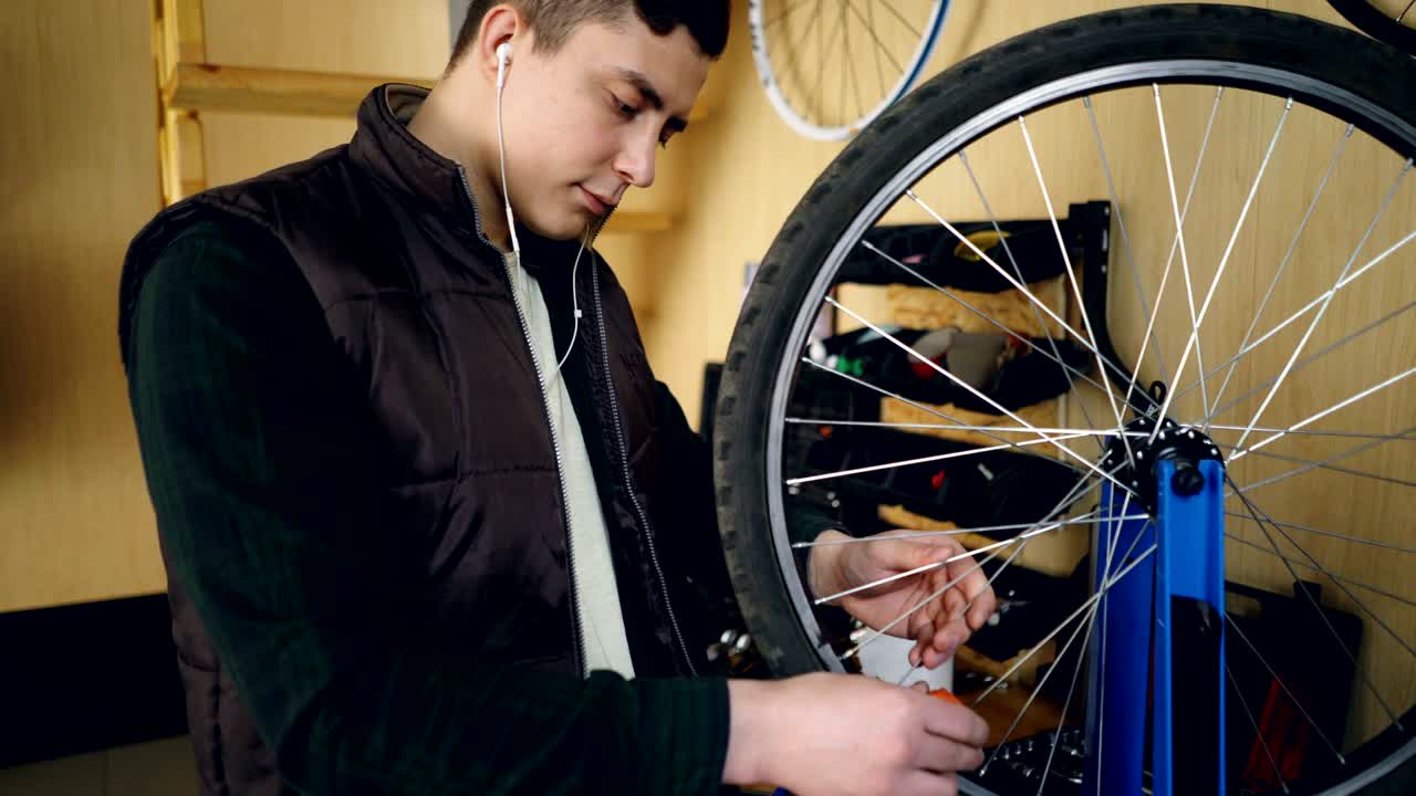 Young man self-employed mechanic is fixing bicycle wheel in workshop. Young man is wearing casual clothes, warm vest and is listening to music with earphones.