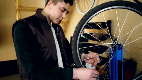 young man self-employed mechanic is fixing bicycle wheel in workshop. young man is wearing casual clothes, warm vest and is listening to music with earphones.