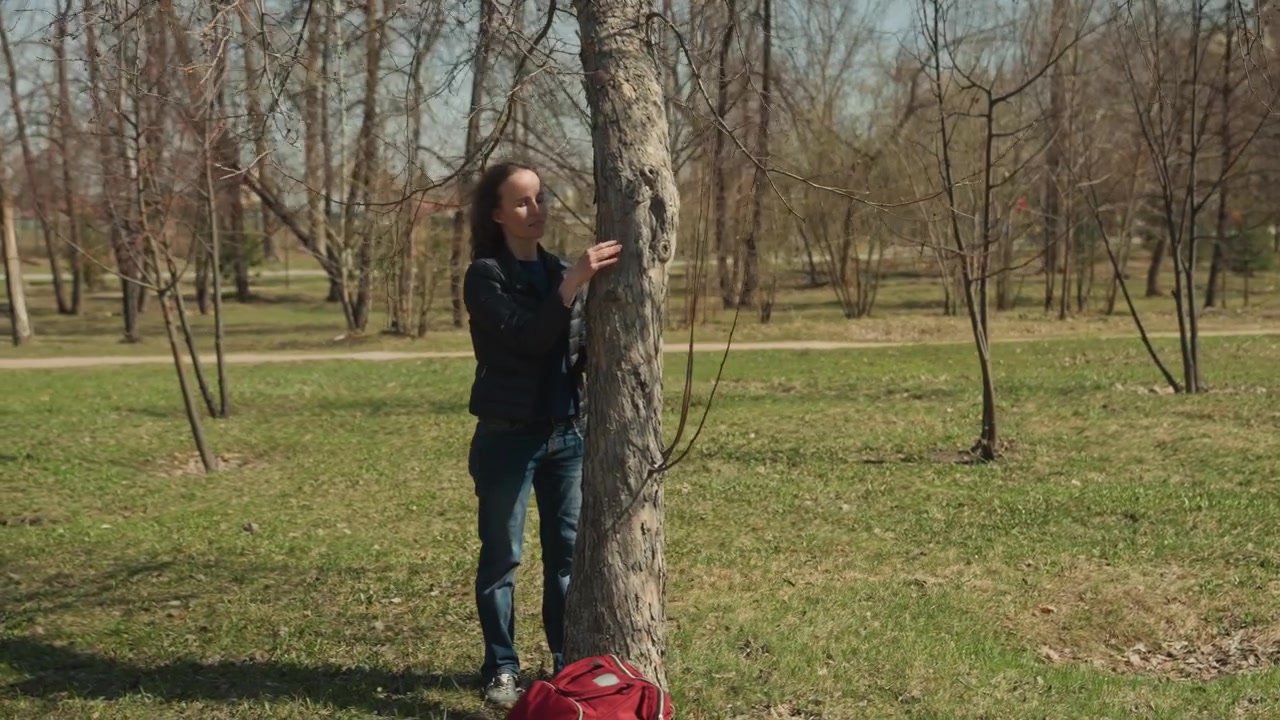 Woman in park closely examining tree bark with hands touching trunk red bag on ground nearby. Outdoor setting with grassy area leafless trees and distant buildings under clear sky