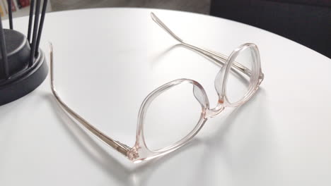 Modern-eyeglasses-in-light-colour-lie-on-a-white-table,-bright-glasses-frame,-slow-motion-footage