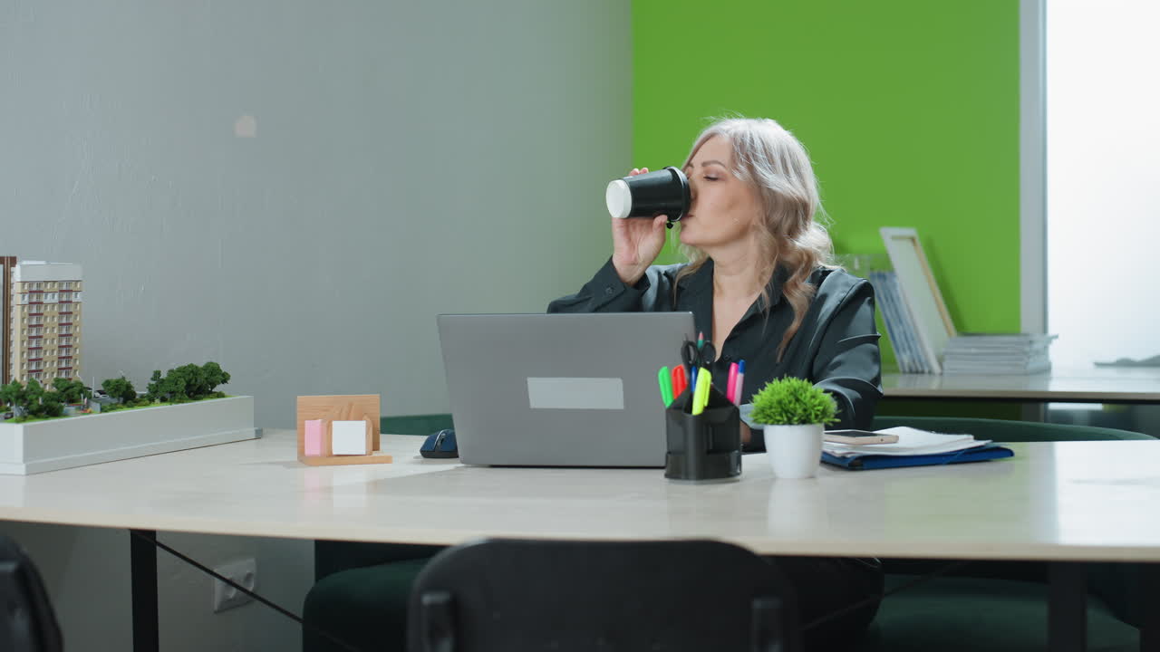 Real estate saleswoman pauses work at office desk to sip coffee before resuming typing on laptop, surrounded by stationery, model building, files, and indoor plants in professional environment