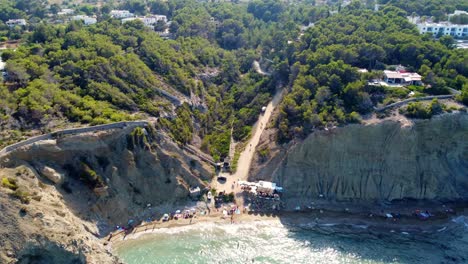 Kleine-Strandbar-Am-Fkk-Hippie-Strand-Auf-Ibiza-Sonnenschirm-Mit-Langem-Schatten-Glatt-Luftaufnahme-Flug-Vorwärts-Fliegen-Drohne-Aufnahmen-Von-Aigües-Blanques-Sommertag-Juli-2022-Marnitz-4k-Filmansicht-Von-Oben