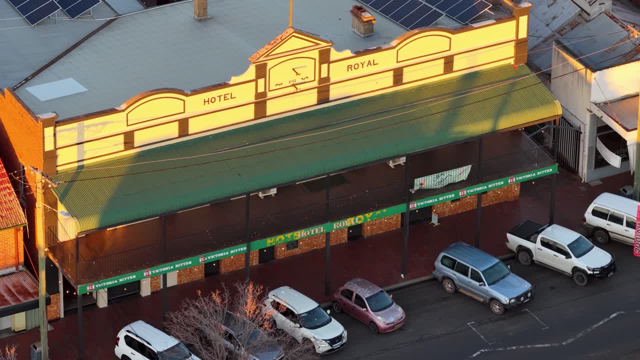 Oblique aerial view of a heritage hotel facade and parked cars in golden hour light, with smooth camera pan revealing street details in Coonabarabran, NSW