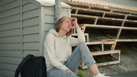 woman in jeans with hand on head, expressing thoughtful reflection while seated in stadium with black bag beside her, background includes worn bleachers