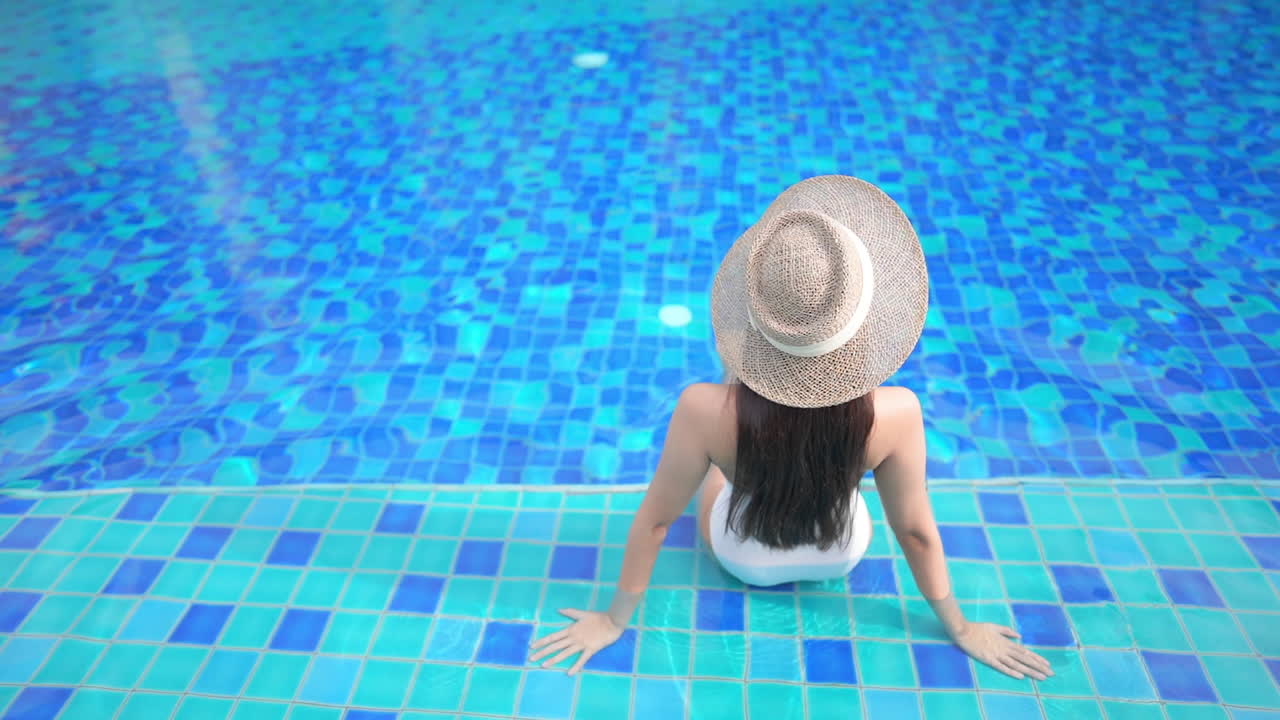 Una donna è seduta sul gradino poco profondo di una piscina, di spalle alla telecamera.