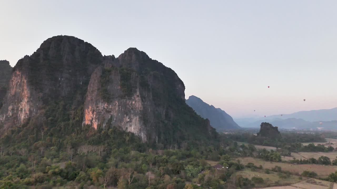 Ripresa aerea con drone delle aspre scogliere calcaree di Vang Vieng all'alba, con mongolfiere che fluttuano sopra il fondovalle nel Laos settentrionale, circondato da una giungla lussureggiante e montagne distanti.