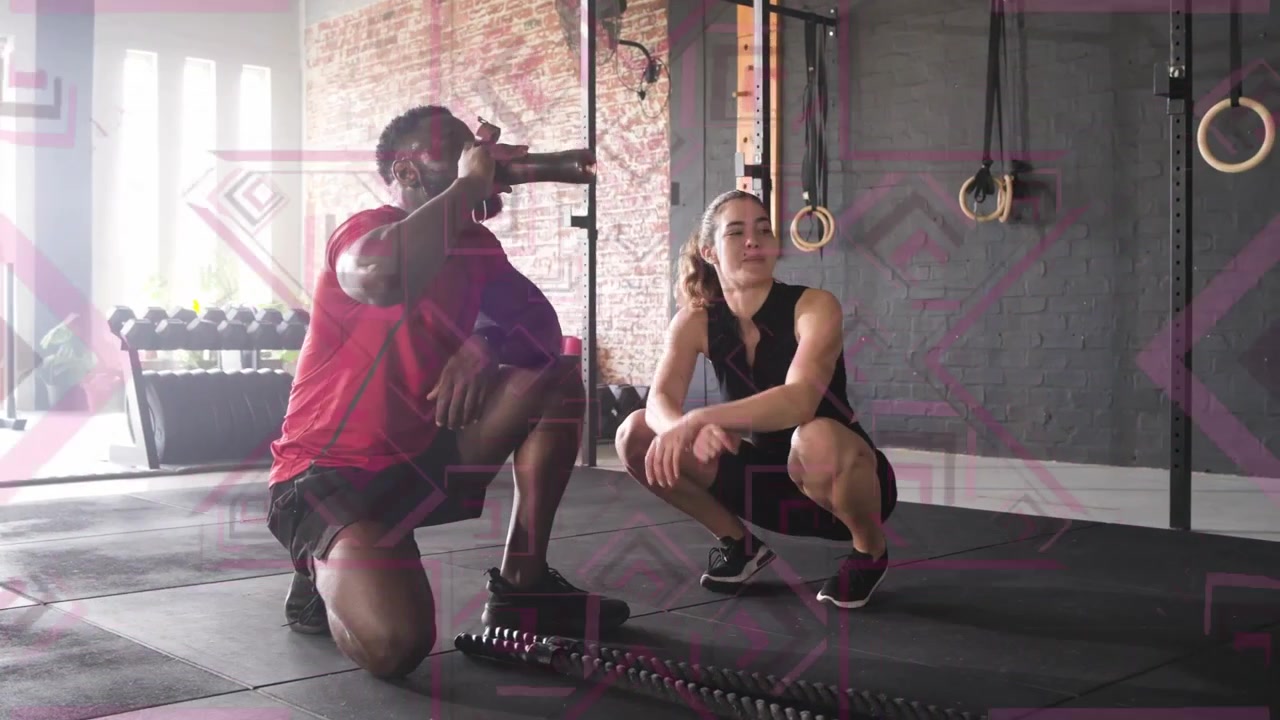 Uomo inginocchiato che sorseggia acqua e poi testa il ginocchio mentre l'allenatore indica le battle ropes e gli anelli in palestra.