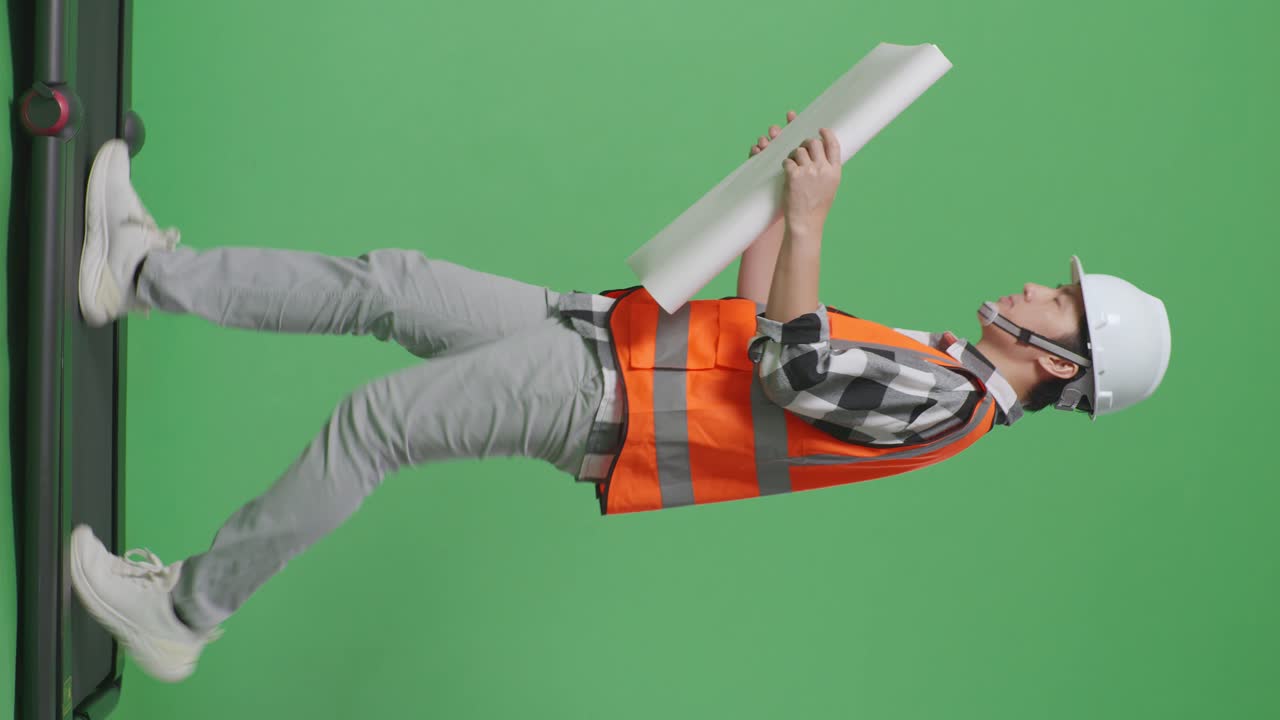 안전 헬을 입은 아시아 남성 엔지니어가 손에 블루프린트를 들고 녹색 스크린 배경 스튜디오에서 는 동안 주위를 둘러보고 있습니다.