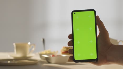 Person-Holding-Green-Screen-Mobile-Phone-With-Traditional-British-Afternoon-Tea-With-Scones-Cream-And-Jam-In-Background