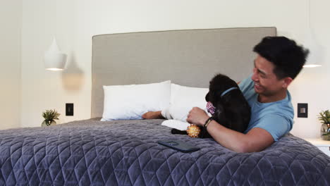 asian man relaxing on bed, joyfully playing with dog and toy at home