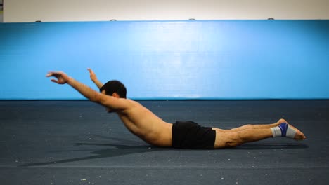 a guy doing an abs workout in a gymnastics gym doing lying down shortless leg and hand raises still shot