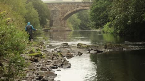 hand-held shot of a flyfisherman walking along a riverbank with a stone bridge