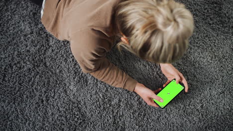Overhead-Shot-Of-Woman-Looking-At-Green-Screen-With-Copy-Space-On-Mobile-Phone-Lying-On-Carpet