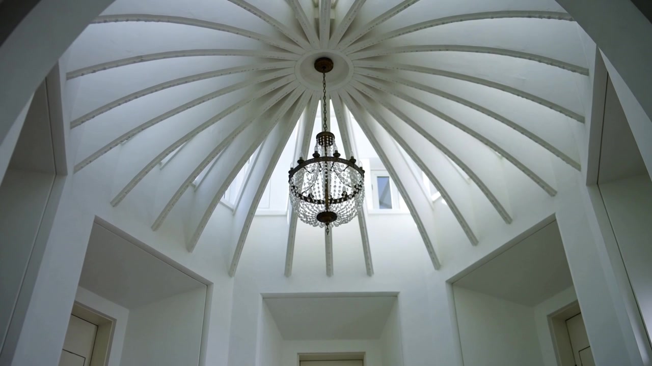 Telecamera che si inclina rivelando un lampadario centrale di cristallo che oscilla sotto una rotonda a cupola, con lucernari.