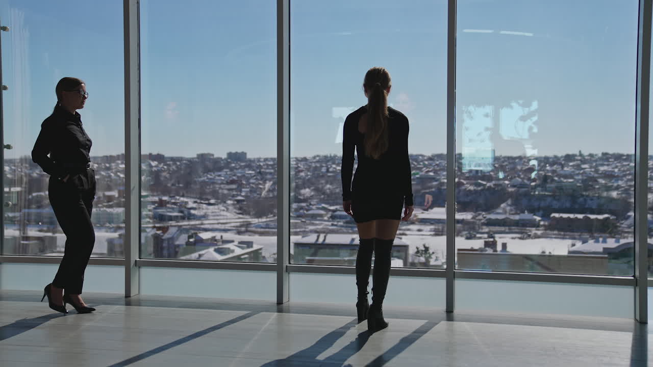 Beautiful lady standing at the window and looking through it. Another lady comes and points at something outside the window.