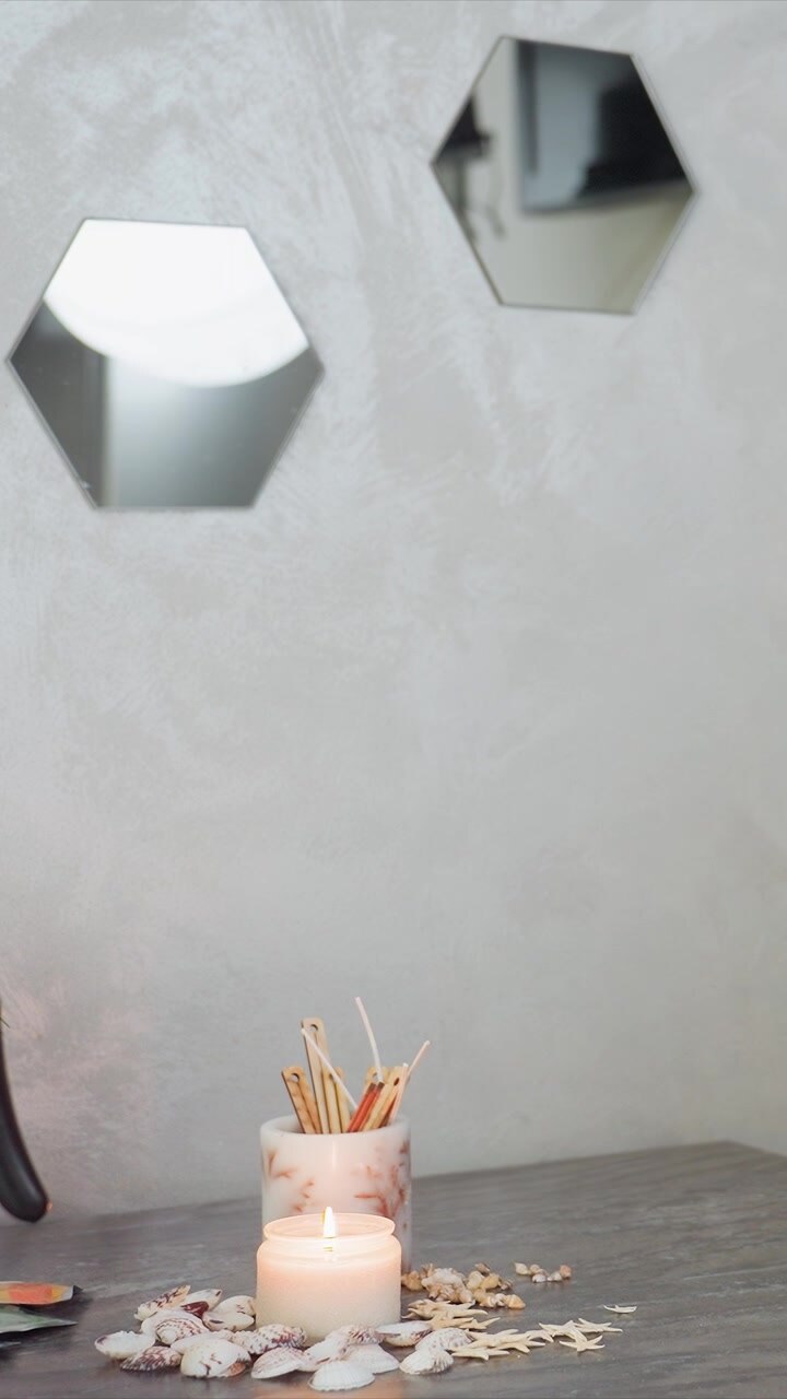 Primo piano delle mani di una donna che tengono vari oggetti su un tavolo, inclusa l'accensione di candele. Una candela accesa in un barattolo di vetro circondata da conchiglie è visibile per tutta la scena.