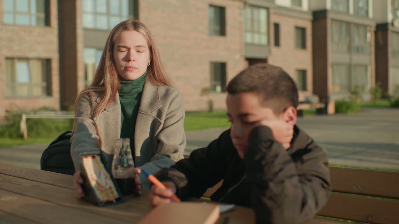 Young boy sits at outdoor wooden table sketching on notebook while woman beside him holds sandwich pack and bottle of juice, both seated near building wall in warm daylight atmosphere