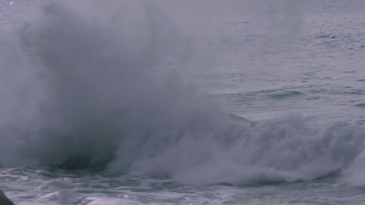 A slow-motion medium shot captures powerful ocean waves crashing against large coastal rocks, with white foam splashing in a dramatic seascape.
