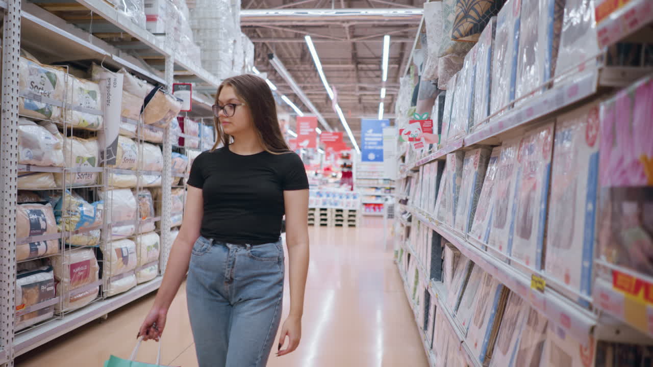 mujer con camiseta negra y vaqueros caminando por el pasillo del supermercado cerca de productos textiles, sosteniendo bolsas de compras y centrándose en las mercancías expuestas en los estantes, entorno minorista brillante