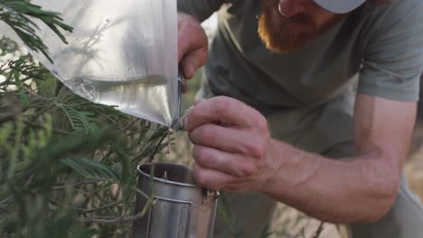 Bearded-caucasian-male-survivalist-gathering-water-from-plastic-bag-on-bush-in-wilderness