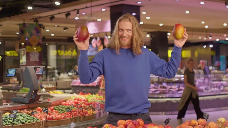 Giovane uomo caucasico che balla con dei mango in mano mentre fa la spesa in un supermercato.