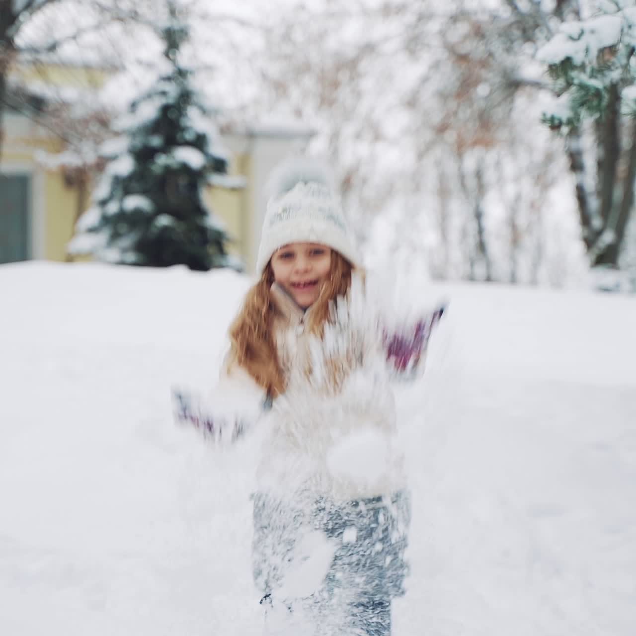 A little girl in warm clothes is descending from the hill and touching the snow. The child is scattering snow by her hands on the background of snowfall in the park. Slow motion