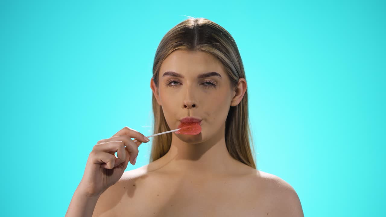 disparo estático de una joven modelo rubia bonita mientras lamia una dulce piruleta en forma de corazón con placer y mira neutralmente a la cámara frente al fondo turquesa