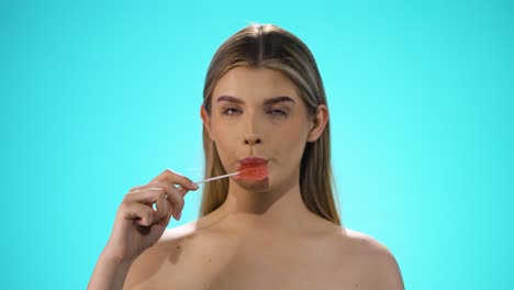 Static shot of a young pretty blonde model while she licks a sweet lollipop in heart shape with pleasure and looks neutrally to the camera in front of turquoise background