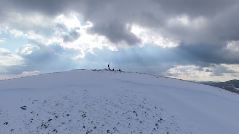 Group-of-hikers-sit-at-snow-covered-round-peak-as-sunlight-rays-stream-across-clouds-and-valley