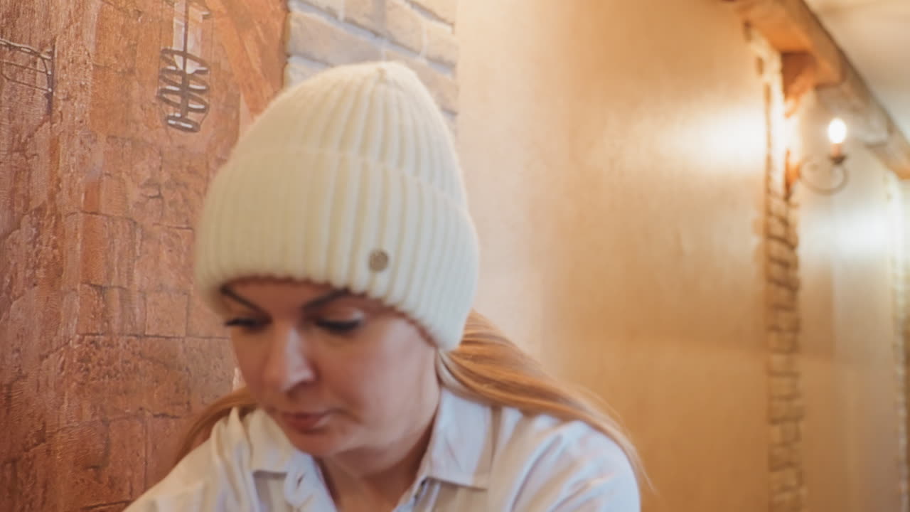 Donna che si sistema il berretto di maglia nel corridoio, tirandosi il cappotto morbido sulle spalle, con espressione concentrata, illuminazione calda sulla parete di mattoni, accogliente ingresso interno, movimenti lenti e deliberati, maglietta neutra, texture morbide