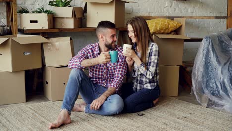 happy married couple clanging mugs and drinking tea while sitting on floor in new apartment after moving in together. relocation, young people and housing concept.
