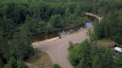 4k-drone-flyby-unrecognizable-man-in-side-by-side-UTV-on-a-river-side-ORV-nature-trail-in-mid-michigan-with-sunny-skies-muddy-water-and-campers-and-trails-all-around