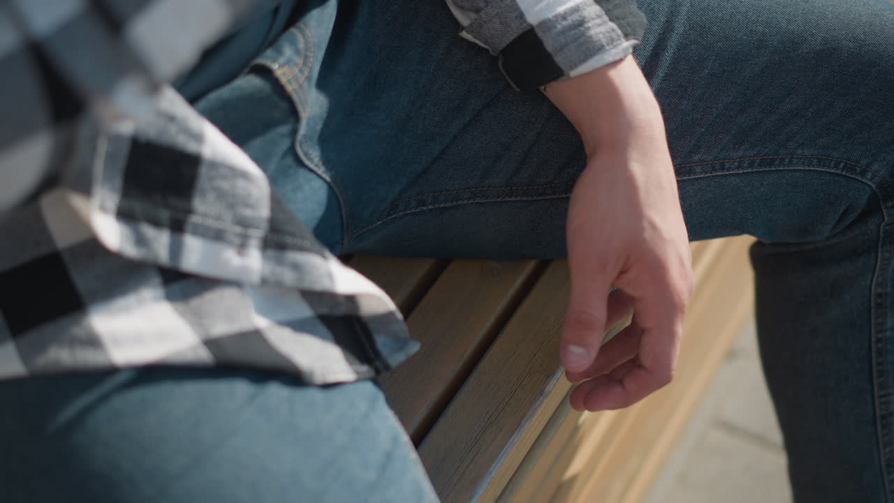 close up of fair skinned student wearing checkered shirt and blue jeans sitting on wooden bench outdoors with relaxed hand resting beside leg after taking red pill medicine on sunny day