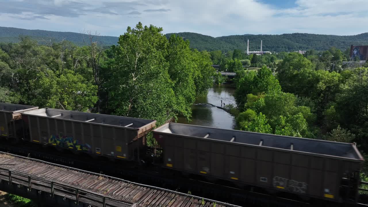 Cargo train with old rusty wagons sprayed with graffiti crossing river in America. Green forest trees and hills of Virginia in distance. aerial view