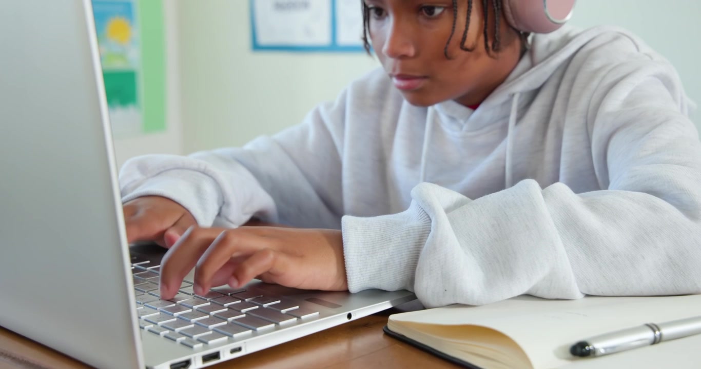 African American teen typing on laptop at desk, wearing pink headphones, using notebook pen