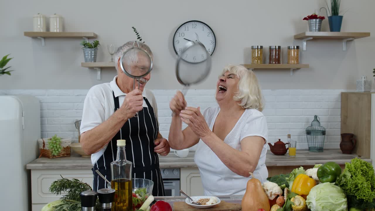 Senior woman and man making a funny dance with strainers. Dancing while cooking together in kitchen