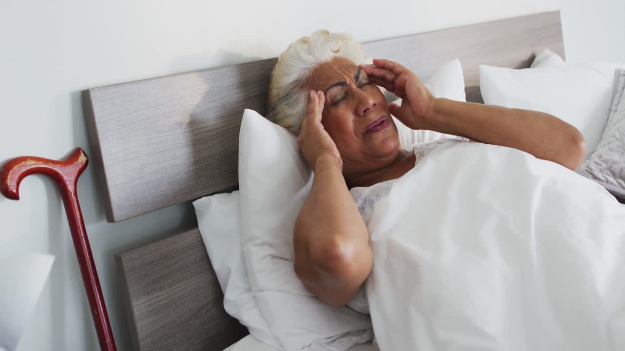 mujer mayor afroamericana enferma sosteniendo su cabeza mientras está acostada en la cama en casa