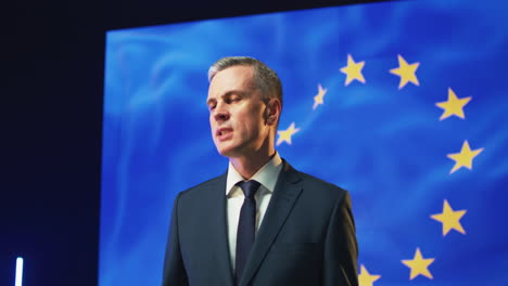 european politician speaking from stage zoom in view of middle aged man in suit speaking about international politics from stage against european stage