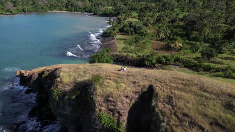 aerial view of two persons on top of grassy ridge cliff overlooking turquoise ocean and tropical coastline, comoros islands