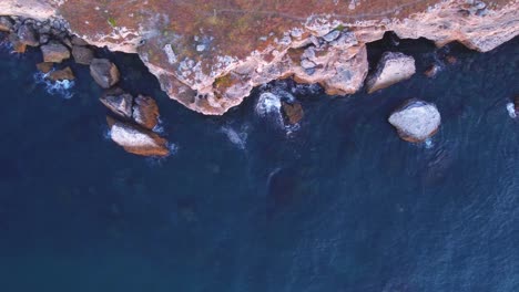 Top-down-aerial-view-of-waves-splash-against-rocky-seashore,-background